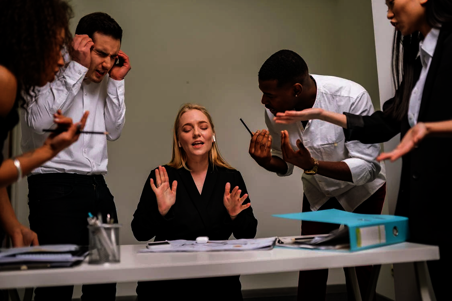 Group of diverse office employees in a heated discussion around a desk, with one woman in the centre appearing calm and trying to mediate workplace conflict.